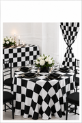 A black and white checkered tablecloth draped over a round dining table, styled with black plates, white floral centerpieces, and black chairs. A matching checkered curtain hangs in the background against a white paneled wall.